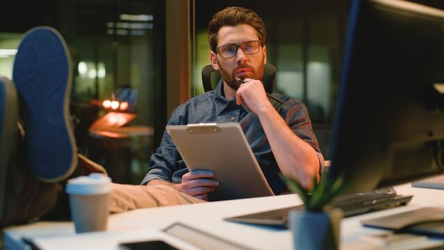 Pensive Caucasian business man thinking idea writing notes looking in computer at night office thoughtful businessman analyst male executive think ponder solution write noting overtime at evening