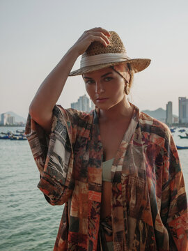 Stylish woman in straw hat and patterned kimono at tropical waterfront with colorful beach lounge