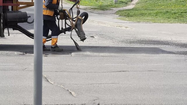 Worker operates specialized road paving machinery, applying hot asphalt mix to repair potholes and cracks on damaged asphalt street surface. Road construction crew performs maintenance.