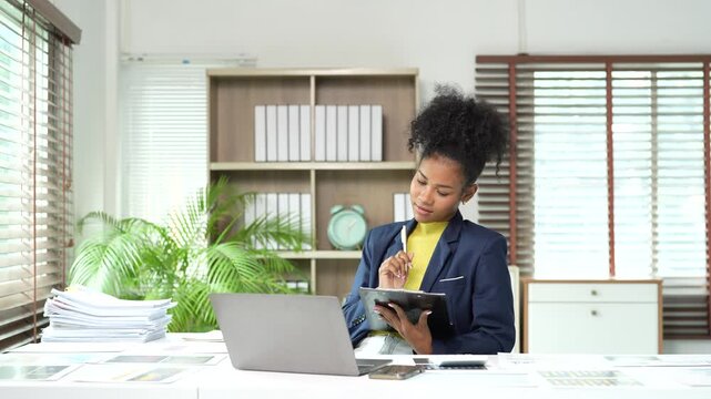 African American businesswoman attending a virtual meeting on her laptop, engaging in a video conference call, and taking notes on a clipboard while sitting at her office desk