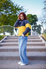 Fototapeta premium Redhead teenage girl model in chunky knit sweater and jeans on stairs background. Bulky knitwear, fashion and style concept