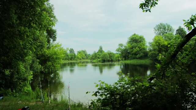 Serene natural landscape of a calm lake reflecting green trees and overcast sky, with multiple fishing rods planted in green grass on left bank, suggesting peaceful outdoor recreation during day