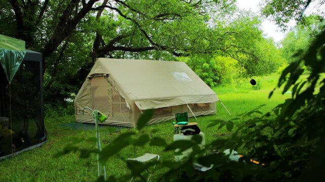 Large beige canvas tent set up in green forest clearing, peaceful summer campsite nestled among lush trees, nobody present, nature outdoor recreation activity