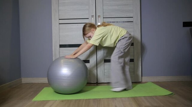 Young Caucasian girl performing fitness exercise using stability ball on yoga mat indoors during daytime, developing core strength and balance at home