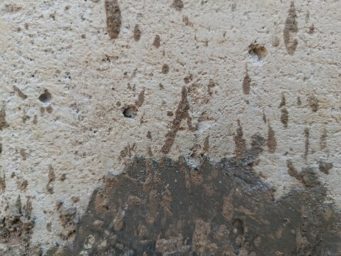 oA close-up shot of a weathered concrete surface featuring a mix of light beige and dark grey textures, characterized by rough patches, small craters, and splattered mud-like markings that create an i