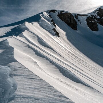 Wind sculpted snow ridge with sharp cornices and exposed dark rock, bright alpine glacier textures under pale winter sky, pristine frozen mountain wilderness in cold sunlight landscape