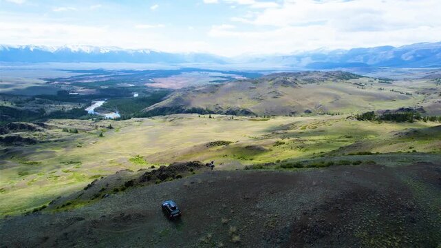 SUV parked on rocky mountain ridge with lone observer facing vast valley, river, snowcapped ranges, layered hills, open sky, and remote scenic wilderness, with landscape, valley, mountain, ridge