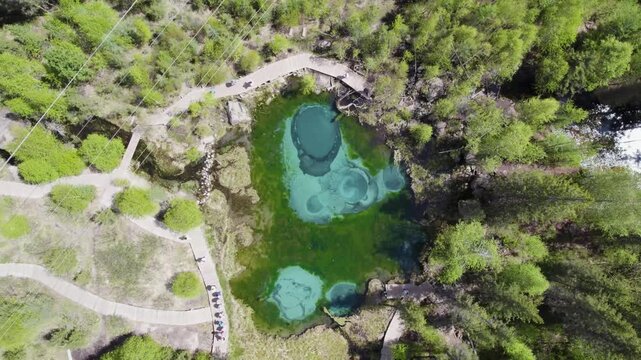 Aerial view revealing turquoise geothermal spring lake with circular mineral rings, curving boardwalks, marshland, forest edge, scenic nature textures, and vibrant thermal colors