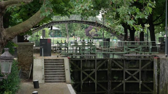 Canal Saint Martin historic lock gates with an arched pedestrian bridge stand surrounded by green trees in Paris France. European water channel infrastructure and transportation history scene.