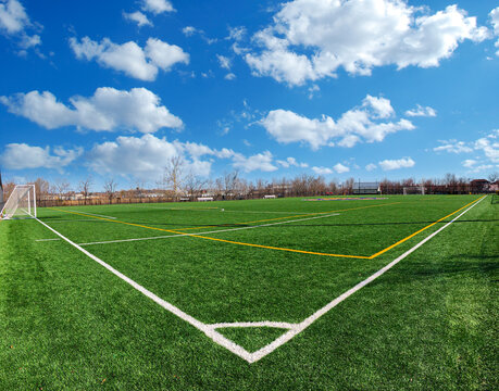 Empty soccer field with green synthetic turf, white markings, and a goal net under a bright blue sky with scattered clouds on a sunny day. Outdoor sports facility, wide angle view.