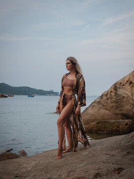 Fashion model in patterned beach kimono and bikini standing on tropical rocky shoreline by calm ocean
