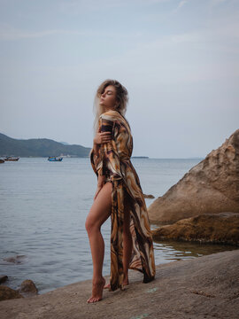 Fashion model wearing abstract print beach kimono posing on rocky tropical shoreline by calm ocean