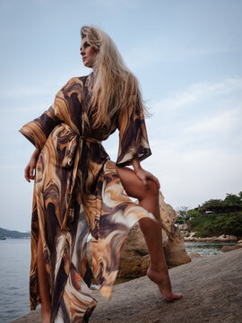 Fashion model wearing abstract print beach kimono posing on rocky tropical shoreline by calm ocean