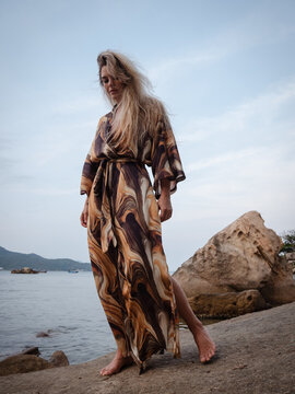 Fashion model wearing abstract print beach kimono posing on rocky tropical shoreline by calm ocean