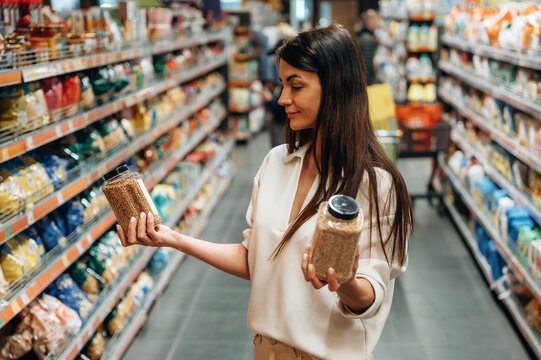 Woman in grocery store is comparing two jars of grains while standing in aisle filled with various food products, showcasing decision-making in shopping experience