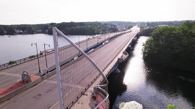Drone view tracking backward across the Kenneth F Burns bridge facing towards Shrewsbury, Massachusetts. Morning sunlight sparkles on the bridge. Car traffic speeds over the bridge in both directions.