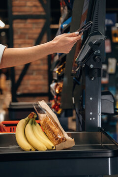 Hand holding smartphone near self-checkout kiosk with bananas and pastry on conveyor, illustrating modern shopping experience and technology integration