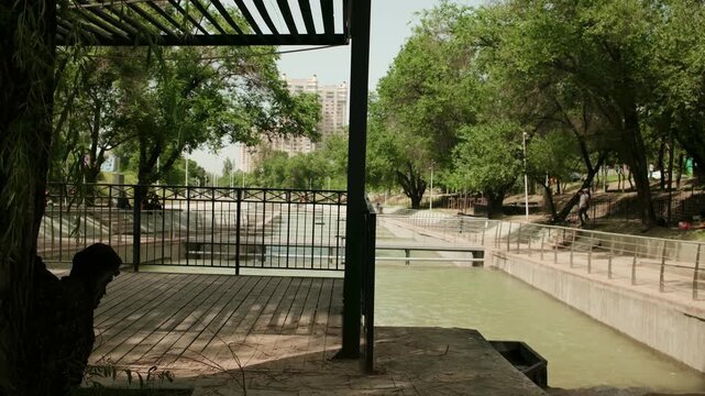 Covered viewing area overlooking a landscaped urban canal in a public park. Modern pergola structure provides shade beside walking paths ano green trees.