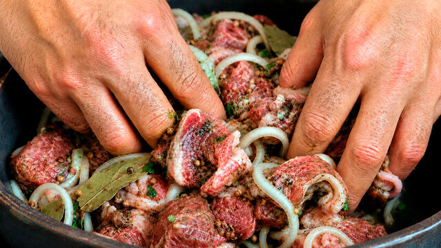 A man's hands mix raw meat with onions and spices in a bowl. Marinating beef pieces with herbs and coriander for a barbecue.