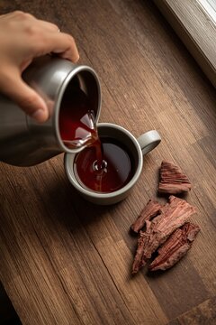 High angle view of dried arjun bark pieces in a ceramic bowl beside a glass mug of dark herbal decoction on a wooden surface.