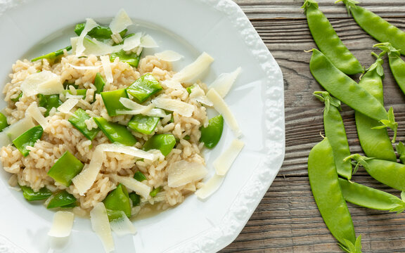 Gourmet Italian risotto with sugar snap peas and parmesan flakes on a white plate with fresh whole pea pods on a rustic wood background.