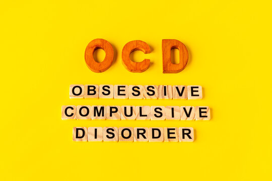 Centered typography layout of OCD abbreviation and Obsessive Compulsive Disorder words spelled with wooden letter tiles on a yellow background.