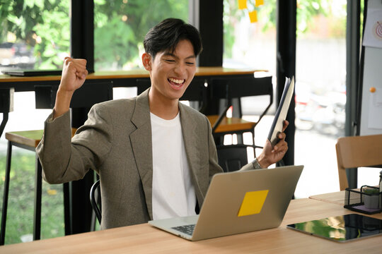 The Job Is Done &ndash; Excited Young Asian Businessman Celebrating Success at Laptop in Modern Office