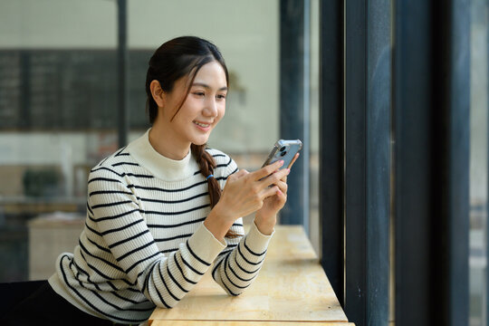 Young Asian Woman Smiling While Using Smartphone by Window in Cafe