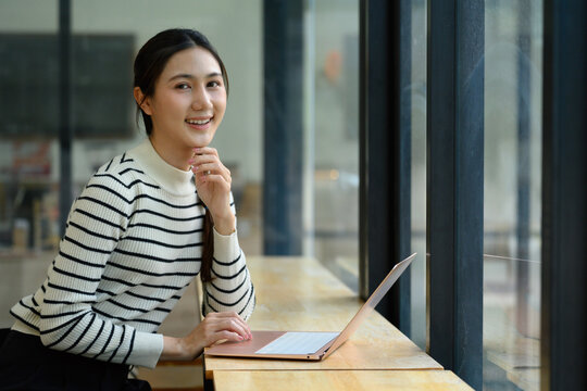 Young Asian Woman Smiling and Thinking While Working on Laptop by Window in Modern Office