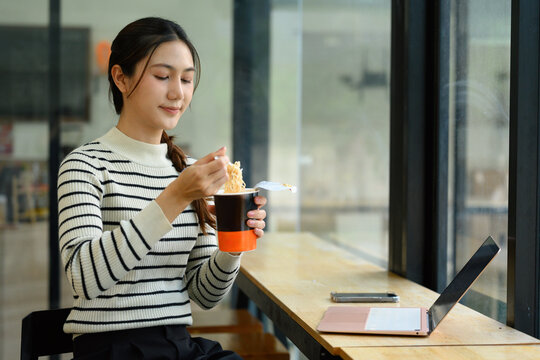 Young Asian Woman Eating Instant Noodles Cup at Office Desk with Laptop During Work Break