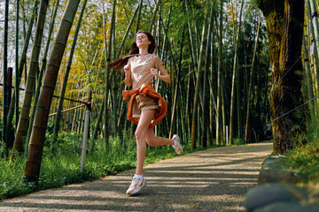 Naklejka premium Woman running along a bamboo forest path, fitness jog and outdoor exercise with a smile, casual shorts and sneakers, sunlight, motion and healthy lifestyle on a scenic trail.