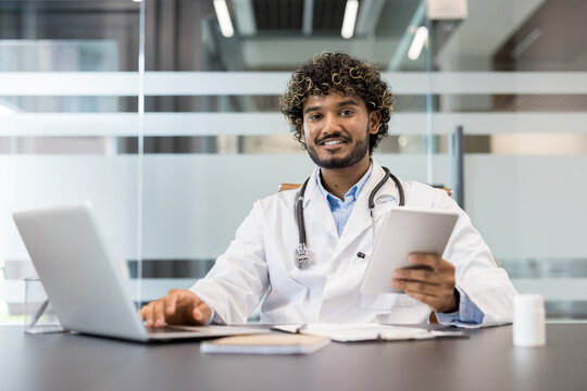 Young male doctor wearing a white lab coat and stethoscope. Sitting at a desk. Using a laptop and holding a digital tablet while looking at the camera and smiling