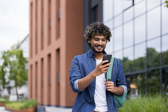 Indian man smiling, wearing a denim shirt and carrying a backpack, actively communicating and navigating online with his mobile phone on a modern university campus setting