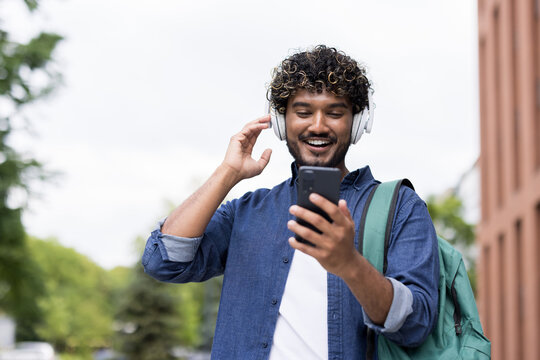 Happy young man with curly hair wearing headphones and a backpack, listening to music or an audiobook on his smartphone outdoors with a joyful expression
