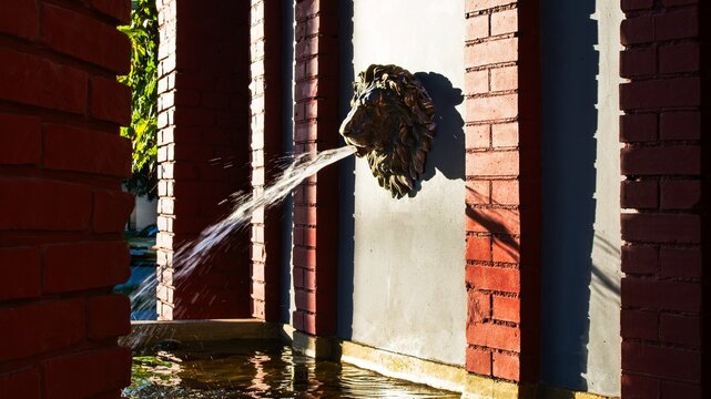 Classic bronze lion head fountain spitting water streams against a red brick column structure. Architectural park design water feature captured in warm sunlight.