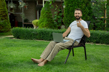 Obraz premium Positive adult man sitting outdoors with a laptop and showing thumbs up gesture. Successful remote work concept, freelance lifestyle, productivity, satisfaction, online business, work from home