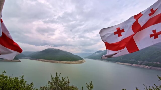 Waving flags of Georgia flutter gently above a calm lake surrounded by misty mountains under a cloudy sky near Tbilisi.