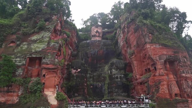 A wide static shot of the colossal Leshan Giant Buddha carved directly into a red sandstone cliff face, with tourist boats crowded at its feet.