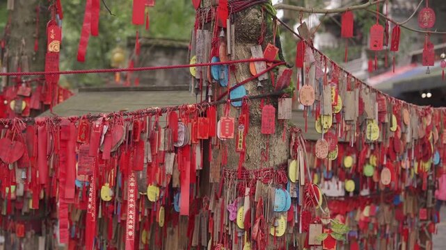 A slow panning shot across thousands of bright red prayer ribbons and wooden wishing plaques hanging from ropes and wrapped around sacred tree trunks in a historic temple courtyard.