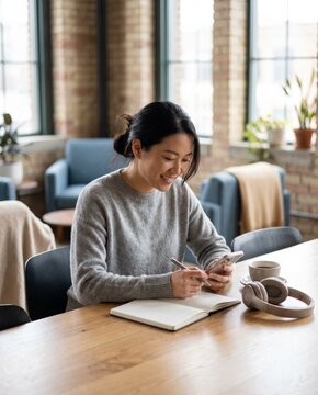 Asian woman using smartphone while taking notes at shared workspace. Concept of mobile productivity, online learning, freelance planning and digital communication.