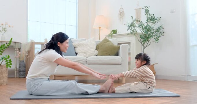 Asian single mother stretches forward on yoga mat facing preschool daughter, guiding simple home exercise with playful encouragement, improving flexibility, concentration, bonding, healthy growth.