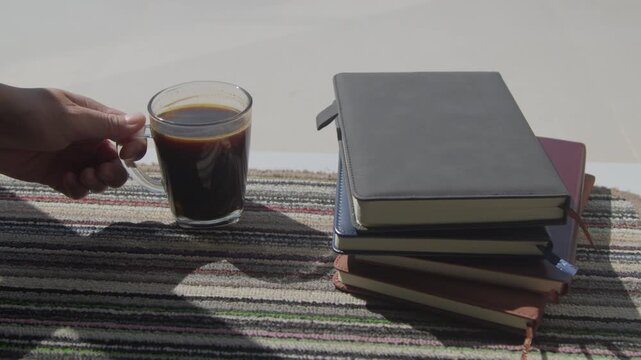 Close-up of hands placing a cup of black coffee next to stacked books on a sunlit carpet, capturing a cozy morning reading and relaxation moment indoors