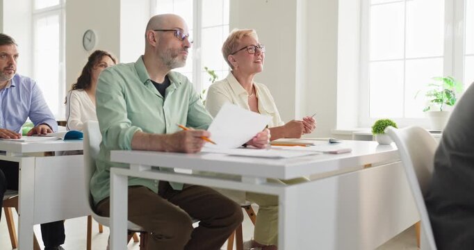 Students seniors adults in classroom learning attentively. Adult learners hold papers and pencils, listening in a bright room. Clear adult learning concept for education marketing needs.