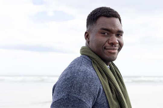 African American man standing on shoreline in blue sweater olive scarf, gazing at ocean, copy space