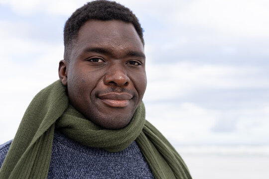 African man standing on shoreline wearing olive-green knit scarf and blue-gray sweater smiling