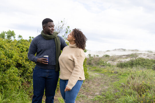 African American couple standing close on coastal dune path wearing sweaters, holding travel mug