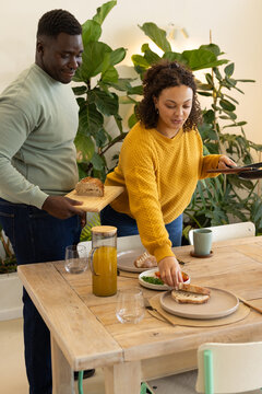 African American couple preparing wooden table, serving bread from cutting board near pitcher