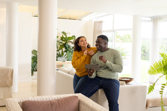 Diverse couple leaning on pale sofa in bright living room, holding tablet, wearing casual sweaters
