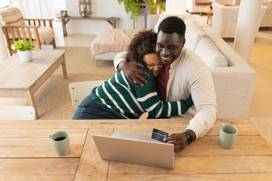 African American couple hugging while sitting at wooden dining table, using laptop and credit card