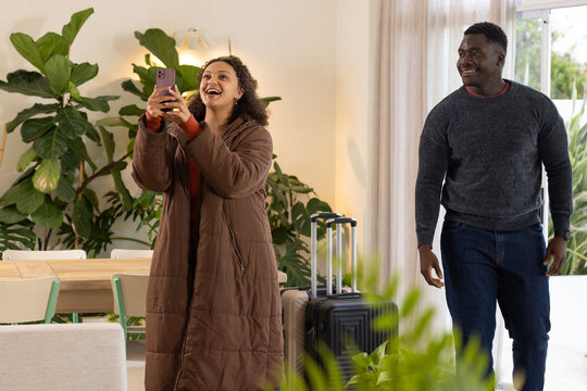African American couple in home, woman holding smartphone wearing brown coat near rolling suitcase